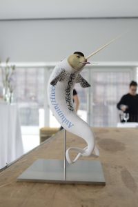 [A photo of a statuette of a bizarre creature that looks like a white narwhal with a fairly anthropomorphic, flesh-coloured face. It is sucking on a lollipop, wearing a black beret, and it has the words “Autumn/Hiver” in decorative text along its side. Its tail fin is shaped like an infinity symbol.]